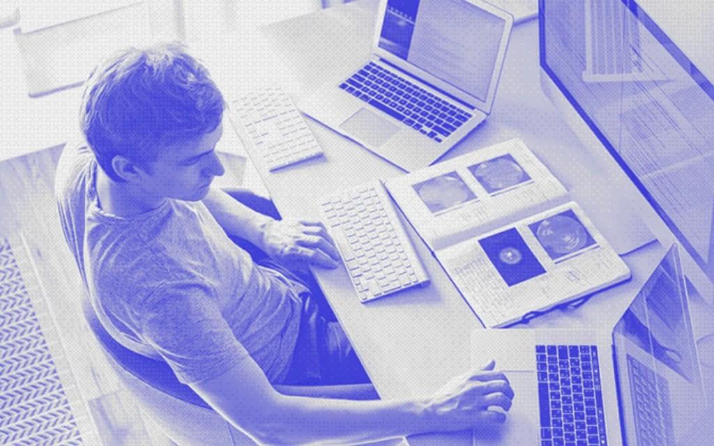 Overhead view of a person at a desk with laptops and monitors, blue duotone halftone style.